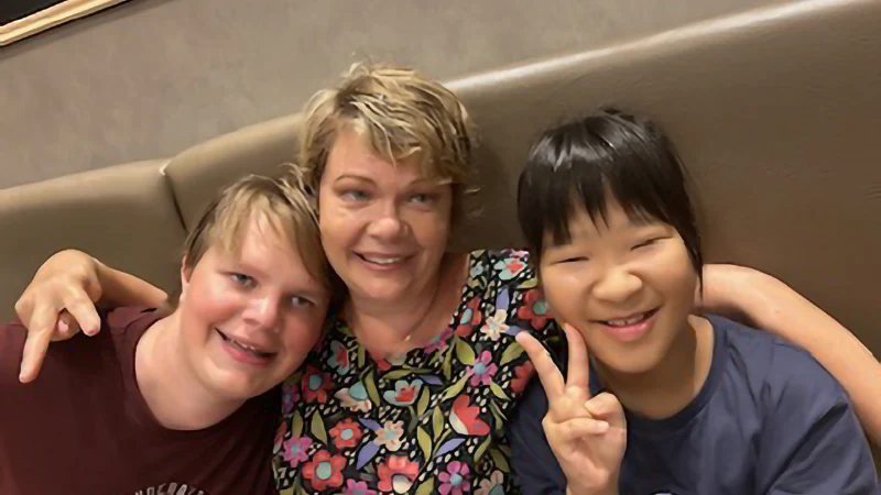 レストランのソファで撮影された外国人家族と日本人女性のセルフィー写真。中央で微笑む短髪の女性を挟んで、左側の男の子と、右側でピースサインをして満面の笑みを浮かべる女の子が写っている仲睦まじい様子。