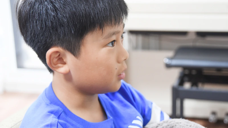 何かをじっと見つめ、思案しているような男の子の横顔のクローズアップ。口元が少し開いており、真剣な表情で話を聞いているか、または考え事をしている様子。背景にはピアノの一部が見える。