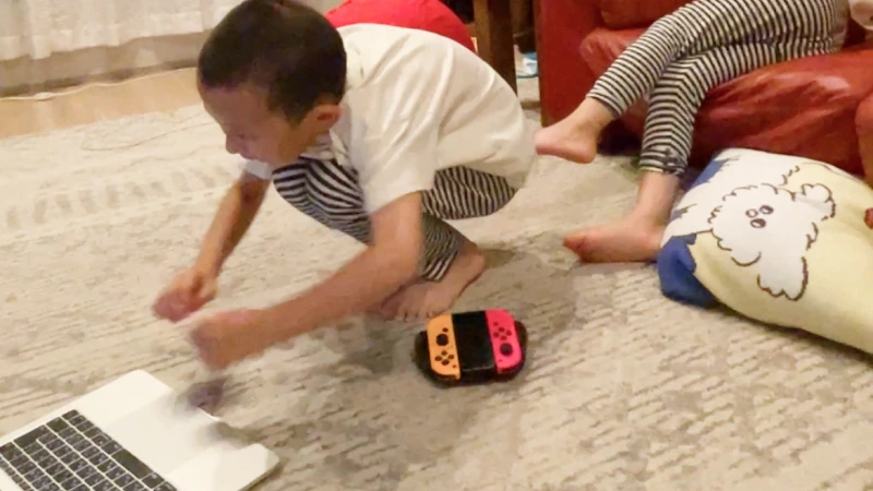 リビングの床に置かれたノートパソコンとNintendo Switchの周りで、男の子が前かがみになって遊んでいる。奥の赤いソファには、男の子と同じボーダー柄のズボンを履いた人物の足が見え、家族とのリラックスした日常のワンシーンであることがわかる。