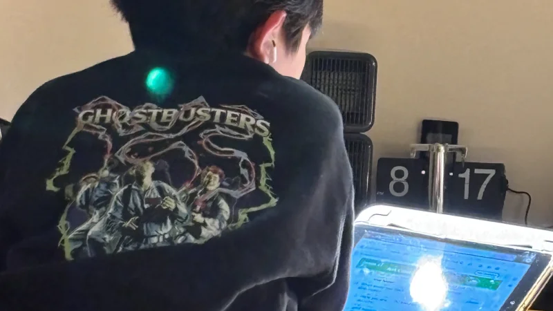 黒い「GHOSTBUSTERS」のTシャツを着た人物の後ろ姿。机に向かってノートパソコンの画面を見ている。画面には青っぽい光が反射しており、オンライン学習か作業をしている様子。右側には「8 17」と表示されたデジタル時計が見える。
