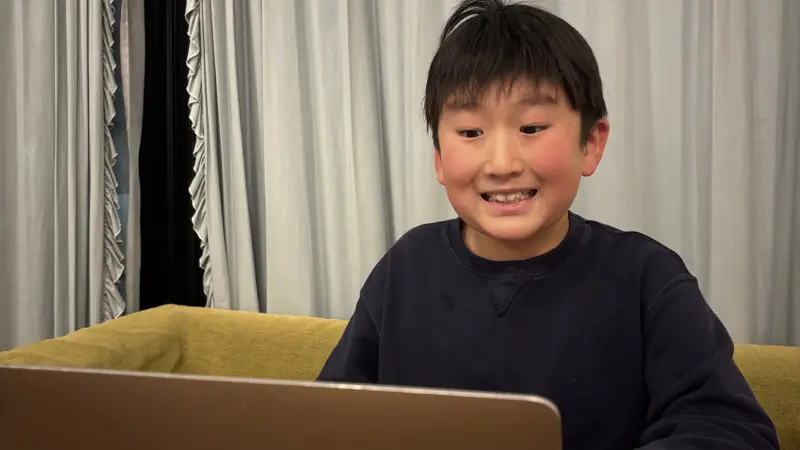 ノートパソコンの画面を見て、嬉しそうな笑顔を浮かべる紺色のセーターを着た男の子。