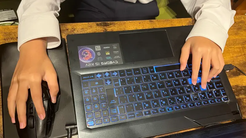 ゲーミングノートPCを使ってゲームをプレイしている手元のクローズアップ。左手は多ボタンマウスを握り、右手は青く光るバックライトキーボードのWASDキー付近を操作している。真剣なゲームプレイの様子が伝わる。