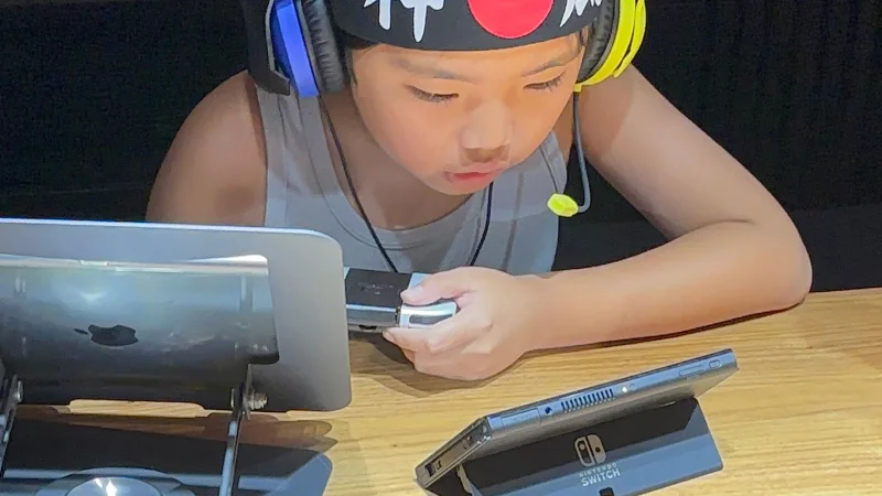 ヘッドセットと「神風」と書かれた鉢巻を付けた男の子が、テーブルの上でNintendo Switchをプレイしている。手元にはJoy-Conを装着した本体を持ち、目の前にはもう一台のSwitchがスタンドに立てかけられている。画面（フレーム外）を真剣に見つめ、ゲームに没頭している。