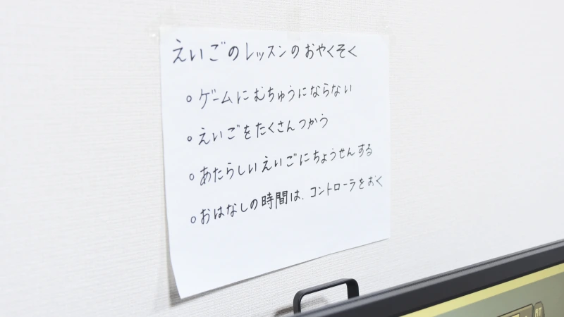 白い壁に貼られた「えいごのレッスンのおやくそく」と題された手書きの張り紙。「ゲームにむちゅうにならない」「えいごをたくさんつかう」「あたらしいえいごにちょうせんする」「おはなしの時間。コントローラをおく」という4つのルールが書かれており、子供が英語学習に真剣に取り組む様子がうかがえる。