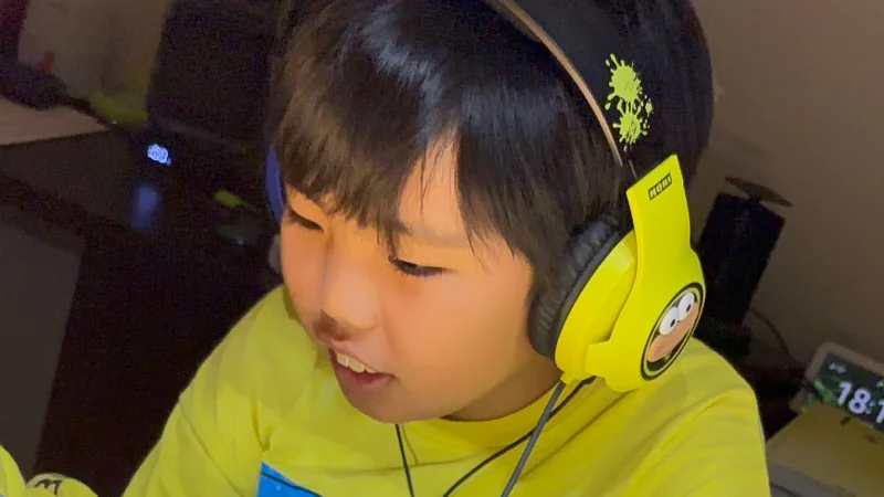 黄色いヘッドセットを装着した男の子の顔のクローズアップ。視線は手元のタブレット（フレーム外）に向けられており、口元が少し緩んで、何かに集中しているか、楽しんでいるような表情。