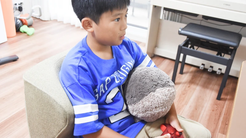 椅子に座り、赤いゲームコントローラーとぬいぐるみを抱えながら、真剣な表情で横を見つめる男の子。青いTシャツを着ており、ゲームに集中しているか、または何か考え事をしている様子がうかがえる。