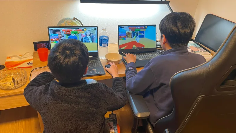 デスクに並んで座り、それぞれノートパソコンでゲーム（ロブロックス）をプレイしている二人の男の子の後ろ姿。