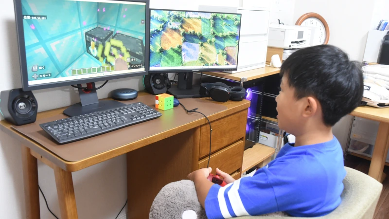 青いTシャツを着た男の子が、椅子に座って2つのモニターが並ぶPCデスクでゲームをしている。両方の画面には「マインクラフト」のゲーム画面が映し出され、手元には赤いコントローラーを握っている。没頭して真剣な表情を浮かべ、広がるゲームの世界を楽しんでいる様子。