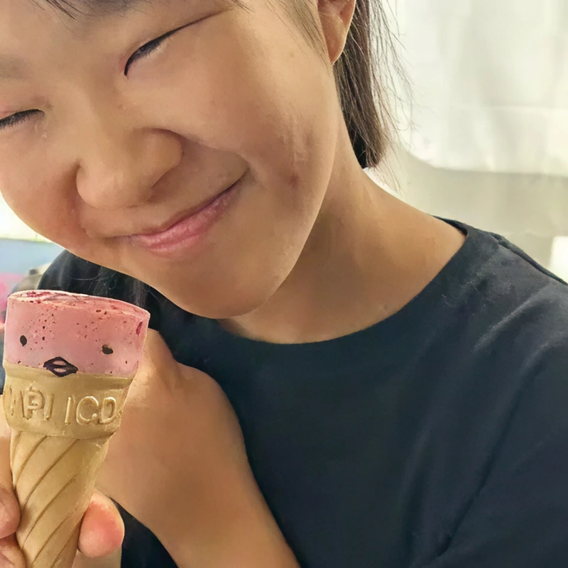 顔のイラストが描かれたピンク色のコーン型チョコレート（カプリコ）を頬に寄せ、目を閉じて幸せそうに微笑む女の子のクローズアップ写真。