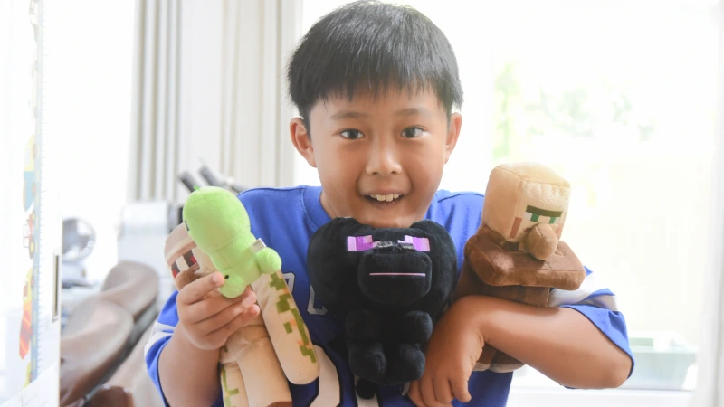 大好きな「マインクラフト」のキャラクターのぬいぐるみを両腕に抱え、にこやかに微笑む男の子。エンダーマン、村人、アイアンゴーレムのぬいぐるみを誇らしげにカメラに見せている。窓からの明るい光が、彼の楽しそうな表情を照らしている。