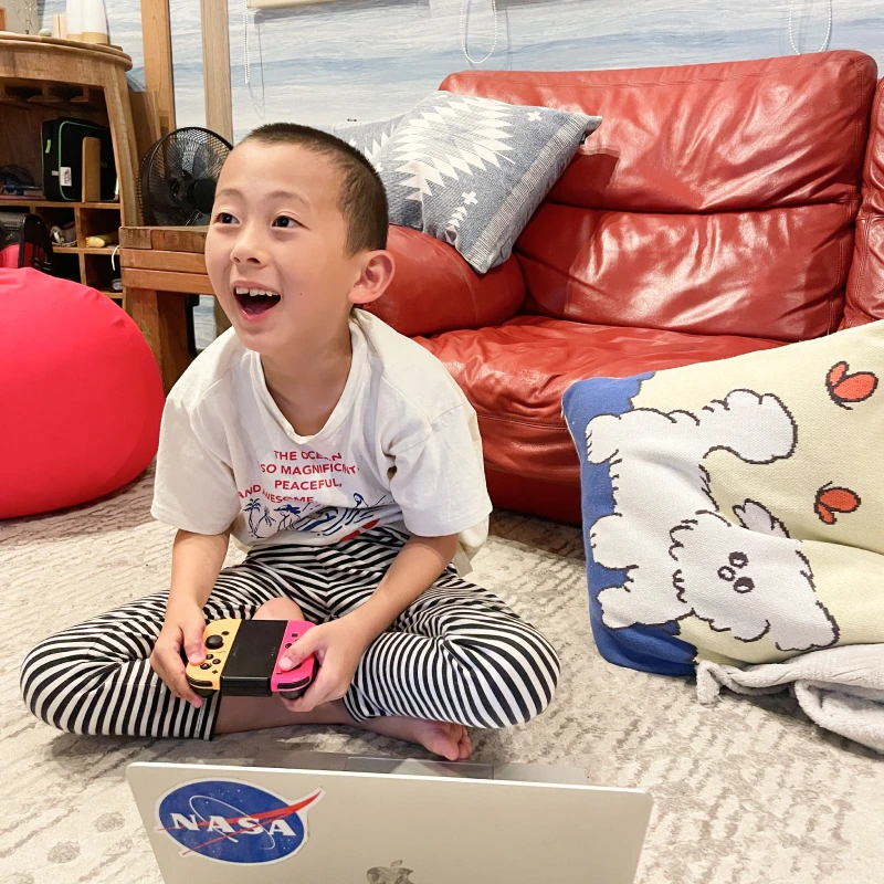 赤いソファの前の床にあぐらをかき、Nintendo Switchを手に持ってゲームに熱中する男の子。とても楽しそうな表情で口を開けて笑っており、ゲームの展開に興奮している様子。目の前にはNASAのステッカーが貼られたノートパソコンが置かれている。