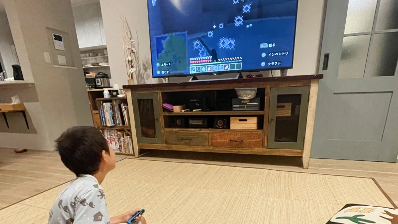 パジャマ姿の男の子が、リビングの床に座ってNintendo Switchのコントローラーを握りしめ、大きなテレビ画面のマインクラフトに熱中している。画面には暗い洞窟を探検する様子が映っており、その世界に没頭する子供の真剣な横顔が見える。