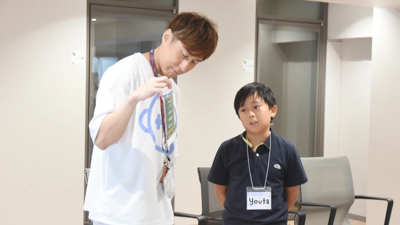 Tシャツ姿のYasコーチと、ポロシャツを着た陽太くんが並んで立っている。コーチはレッスルールが書かれた紙を持っており、子供は「Youta」と書かれた名札を胸に、真剣な表情でそのルールについて説明をしている。