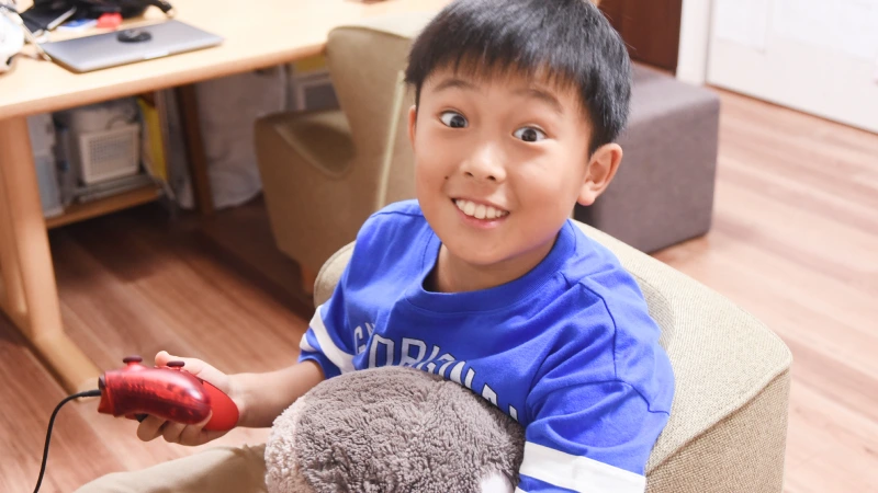 椅子に座り、赤いゲームコントローラーを片手に持ち、カメラに向かっておどけた表情を見せる男の子。少し目を大きく見開いて笑顔を作っており、遊び心いっぱいの瞬間を切り取った一枚。