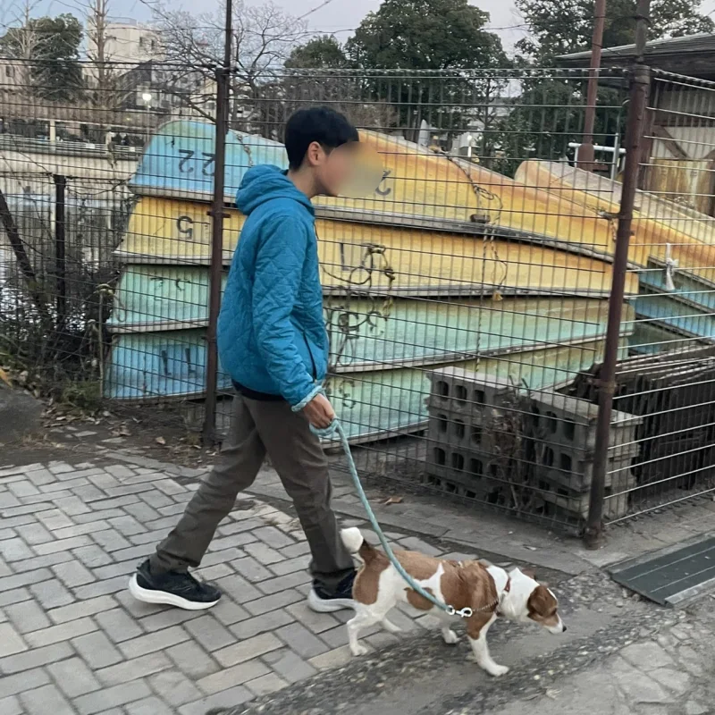 青いジャケットを着た男の子が、フェンス沿いの歩道で犬を散歩させている様子。フェンスの奥にはカラフルなボートが積まれている。
