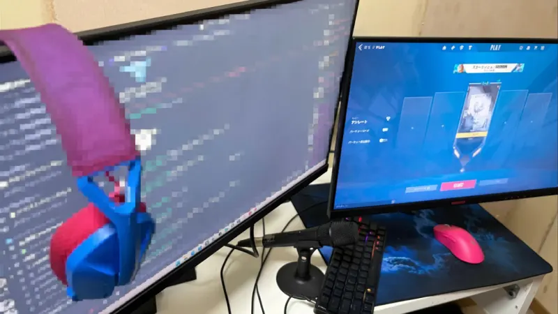 デュアルモニターのPCデスク環境。左側のモニターにはDiscordの画面（モザイク処理あり）が映り、縁に青とピンクのヘッドホンが掛けられている。右側のモニターにはゲーム「ヴァロラント（VALORANT）」のプレイ画面が表示されている。デスク上にはスタンドマイク、キーボード、青いマウスパッド、ピンク色のゲーミングマウスが置かれている。