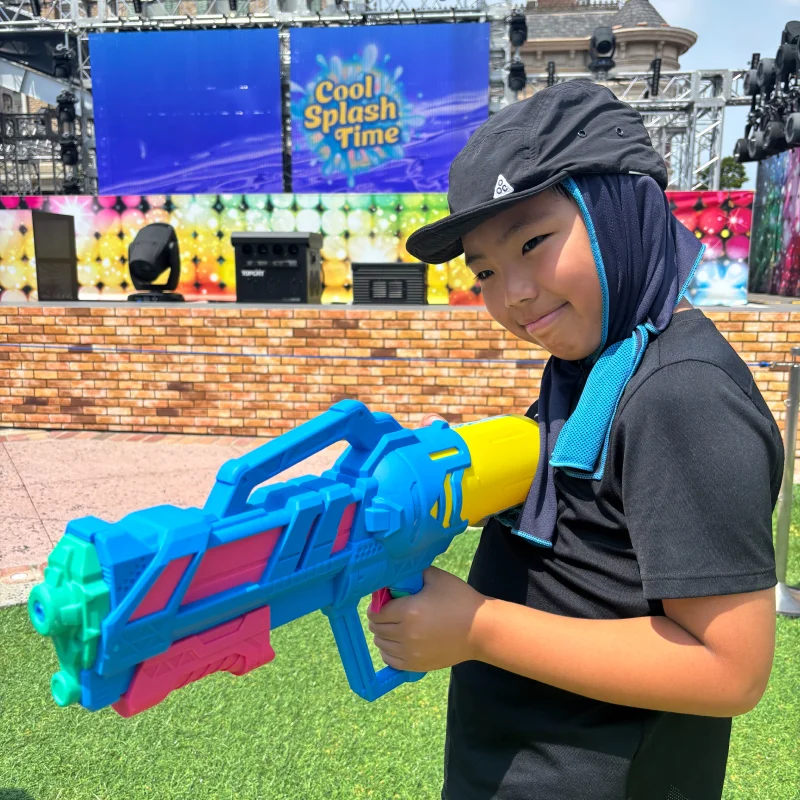 屋外のイベント会場で、日除け付きの帽子とタオルを身につけた男の子が、カラフルで巨大な水鉄砲を構えてポーズをとっている。背景には「Cool Splash Time」と書かれたステージがあり、夏のイベントを楽しんでいる様子で、少しはにかんだような笑顔を見せている。