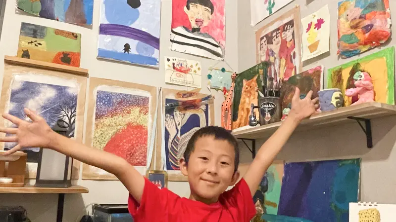 自分の作品である絵画で埋め尽くされた部屋の中で、赤いTシャツを着た男の子が、胸を張って誇らしげに両手を広げている。壁や棚には、彼が描いたカラフルな作品が隙間なく飾られており、子供の創造性と喜びに満ちた空間であることが伝わってくる。