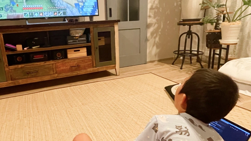 猫の柄がプリントされたパジャマを着た男の子が、リビングの床に座ってテレビゲームに集中している。隣にノートパソコンを開いたまま、大きな画面に映る「マインクラフト」の世界を冒険している様子。
