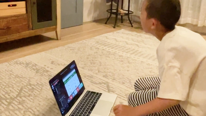 ボーダー柄のズボンを履いた男の子が、リビングの床に座ってノートパソコンを前にしている。画面にはオンライン学習かビデオ通話の様子が映し出されているが、男の子は少し視線を上げ、前方を真剣に見つめている。