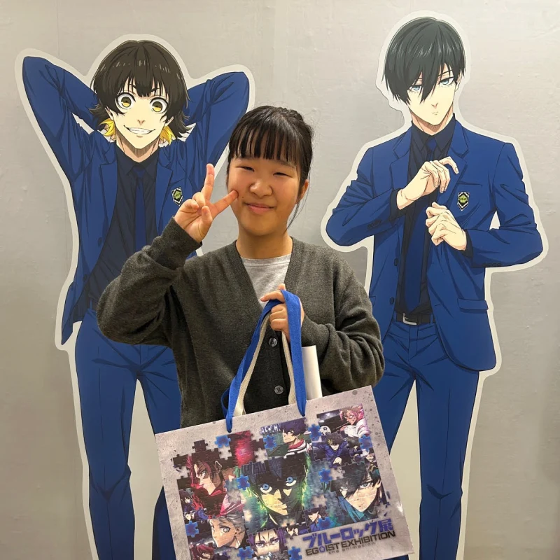 アニメ「ブルーロック」のキャラクター（蜂楽廻と糸師凛）の等身大パネルの前で、女の子がピースサインをして笑顔で立っている。手には同アニメのイラストが描かれたショッパーバッグを持っており、イベントや展示会を訪れた記念写真のようだ。
