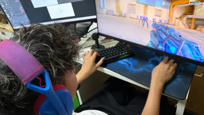 青とピンクのヘッドホンを装着し、PCゲーム「ヴァロラント（VALORANT）」をプレイしている人物の後ろ姿。左手でキーボード、右手でマウスを操作しており、ゲームに集中している様子がうかがえる。左側のサブモニターにはDiscordの通話画面が開かれている。