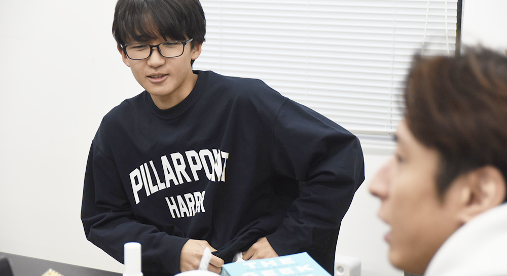 黒いスウェットシャツと黒縁の眼鏡をかけた若い男性一宇くんが、少し身を乗り出し、真剣な表情で話している。右側にはYasコーチの横顔が写り込んでおり、インタビューで会話している場面だとわかる。