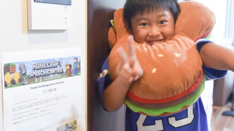 ハンバーガーの形をしたユニークなネックピローを首にかけた男の子が、満面の笑みでピースサインをしている。隣の壁には「MINECRAFT SPEECH CONTEST」と書かれた賞状が貼られており、コンテストでの頑張りを喜ぶ、遊び心にあふれた一枚。