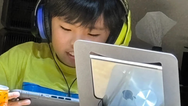 黄色いTシャツを着た男の子が、銀色のスタンドに立てかけられたタブレット端末を操作している手元のクローズアップ。左手にはお菓子のようなものを持っており、リラックスして画面に見入っている様子。