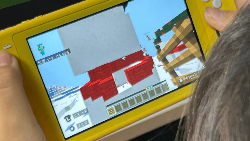 黄色のNintendo Switch Liteの画面のクローズアップ。マインクラフトのゲームプレイ画面が表示されており、プレイヤーが赤と白のブロックを使って何かを建築している様子が写っている。