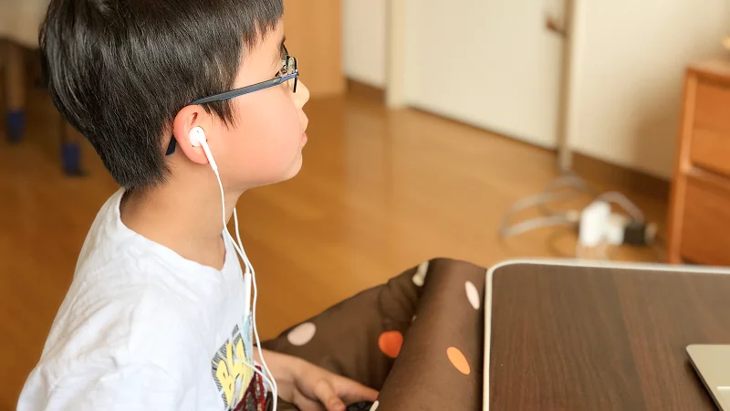 眼鏡をかけた男の子が、こたつに入って白いイヤホンを耳にし、机の上の画面をじっと見つめている横顔。eスポーツ英会話をテストプレイしている様子で、真剣に集中しているのがわかる。