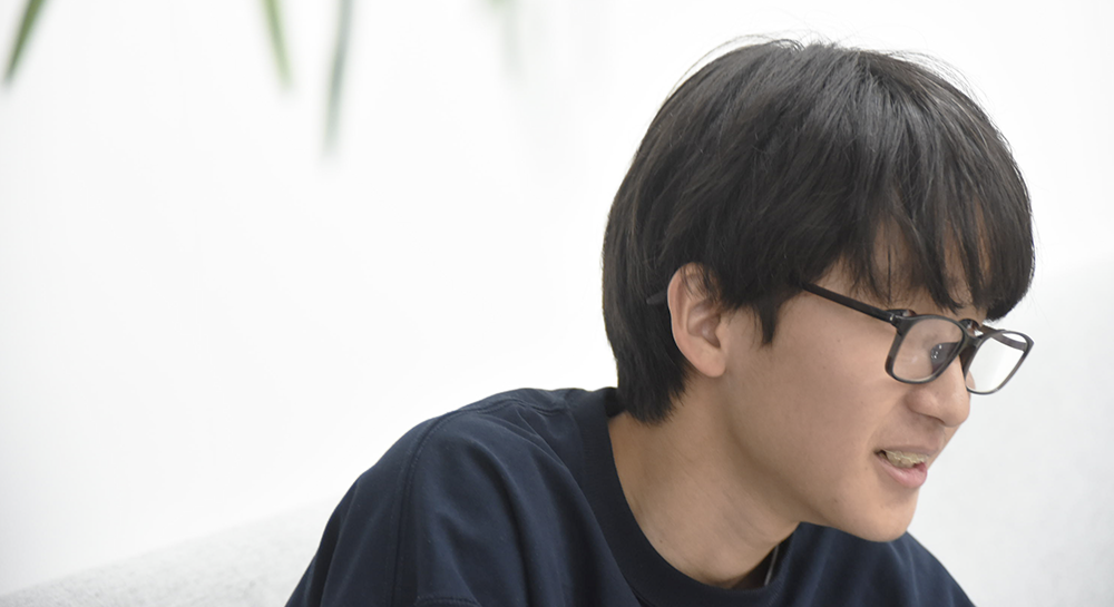 インタビュー中に、少しうつむき加減で穏やかに話す男性のクローズアップ。黒縁の眼鏡をかけ、何かを丁寧に説明しているような真剣な表情が印象的。