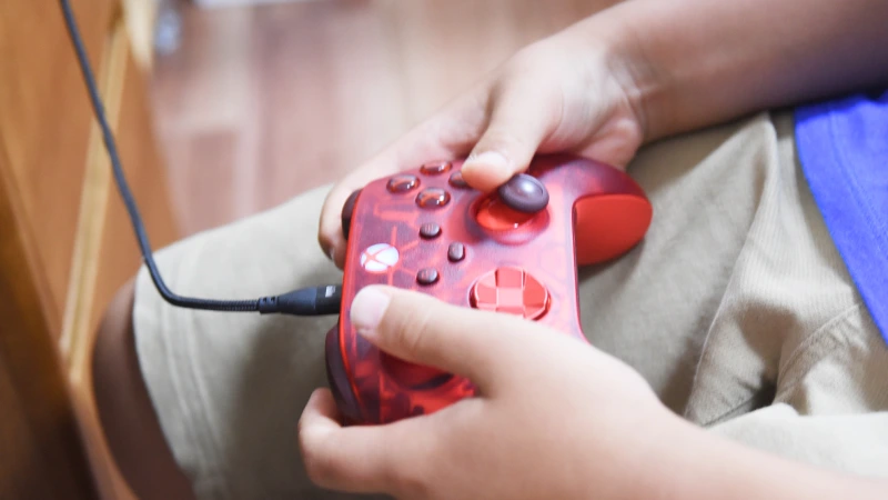 子供が両手で赤いスケルトンデザインのゲームコントローラーを握り、真剣に操作している手元のクローズアップ。指がボタンやスティックを素早く動かしており、ゲームへの集中ぶりがうかがえる。