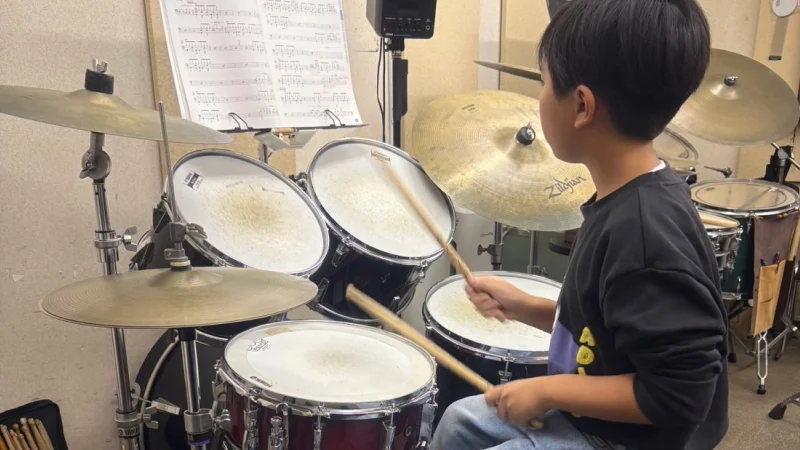 スタジオでドラムセットに向かって座り、演奏している男の子の後ろ姿。譜面台に置かれた楽譜を見ながら、右手のスティックでシンバルを叩こうとしている瞬間。真剣に練習に取り組んでいる様子がうかがえる。
