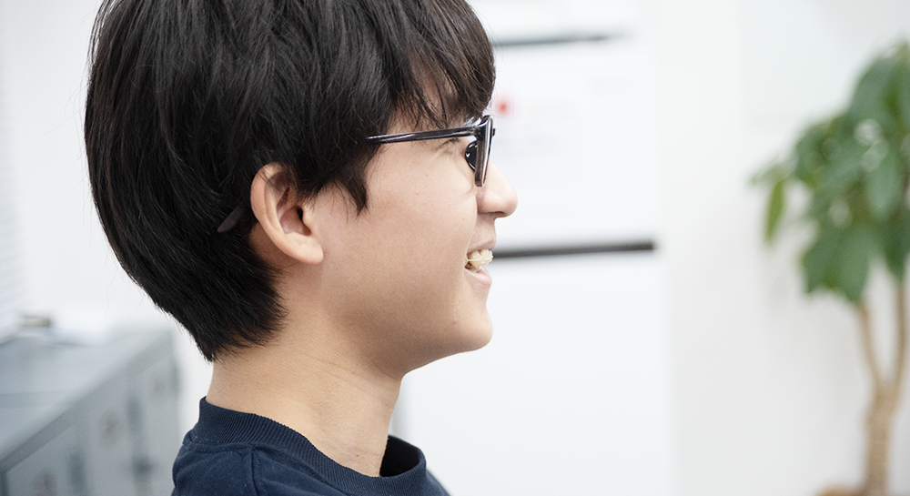 インタビュー中に、相手の話を聞いて微笑んでいる男性の横顔のクローズアップ。黒縁の眼鏡をかけており、和やかな雰囲気で対談が進んでいることが伝わる。背景には観葉植物が見える。