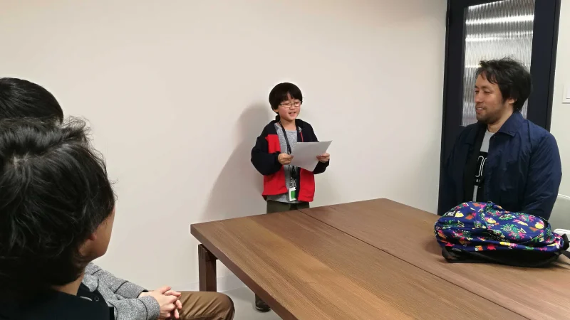 会議室のような部屋で、数人の大人の前で一生懸命に発表している男の子。赤い上着を着た男の子は、手元の紙を読み上げており、隣に座る男性や手前にいる参加者は温かい表情でその様子を見守っている。