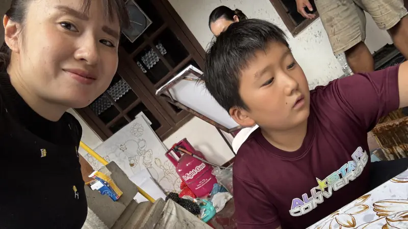 自撮りのアングルで、黒い服を着て微笑む女性と、その隣で花柄の描かれた大きな白い紙（または布）に向かって熱心に何かを描いている（または色を塗っている）男の子の様子。男の子はマルーン色の「ALL STAR CONVERSE」のTシャツを着ており、下を向いて作業に集中している。背景には Bright Gas と書かれたピンク色のガスボンベや、他の人の足が見える。