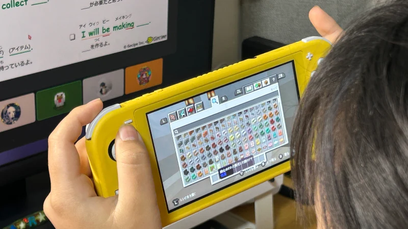 黄色のNintendo Switch Liteでマインクラフトのインベントリ画面を開いている女の子の手元（肩越し視点）。奥のPCモニターには「I will be making...」などの英文が表示された学習用画面が見え、ゲームと学習を並行して行っている様子がうかがえる。