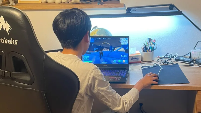 黒いゲーミングチェアに座り、デスク上のノートパソコンで「フォートナイト」をプレイしている男の子の後ろ姿。右手でマウスを操作している。
