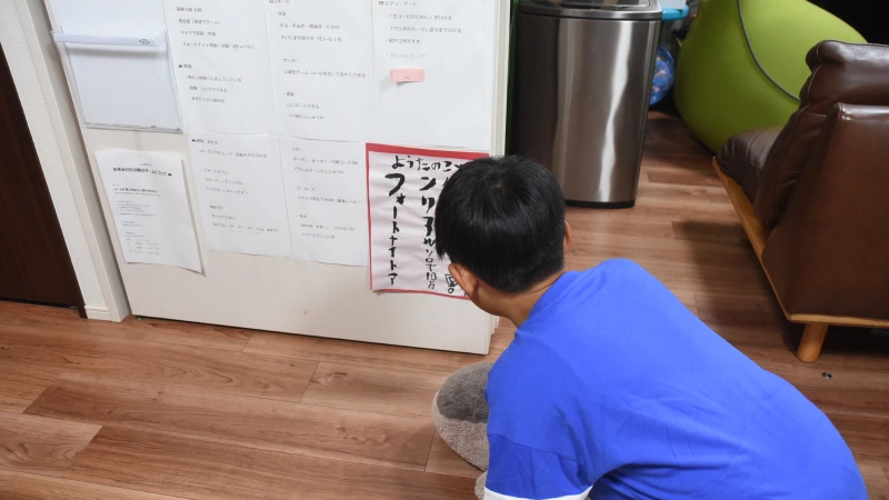 青いTシャツを着た男の子が、床に座って壁に貼られた紙を真剣な表情で見つめている。紙には「ようたのことしの目標 フォートナイトでアンリアルをソロで目指す」という手書きの文字が大きく書かれており、それを説明してくれているシーン。
