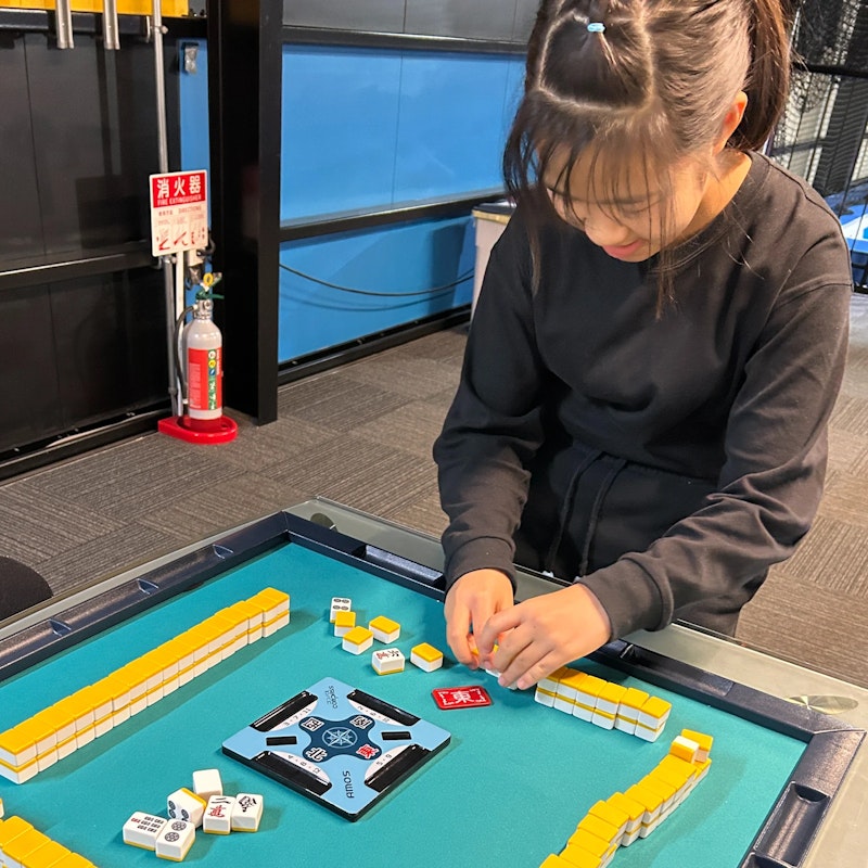 黒い長袖シャツを着た女の子が、全自動麻雀卓に向かって牌を揃えている様子。真剣な手つきでゲームの準備または片付けをしており、背景には消火器が見える。