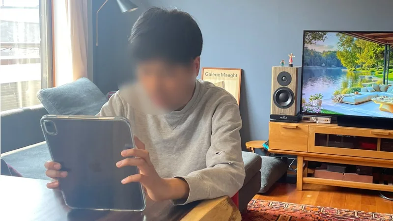 リビングのテーブルに座り、両手でタブレット端末を持って画面を見ている男の子。背景にはテレビや木製の家具がある。