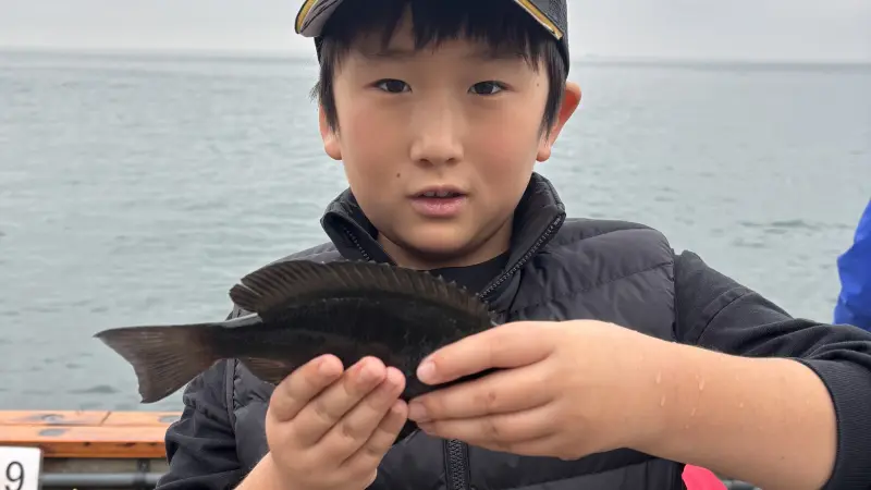 海に浮かぶ船の上で、自分が釣り上げた黒い魚を両手でしっかりと持ち、誇らしげに見せている帽子を被った男の子。