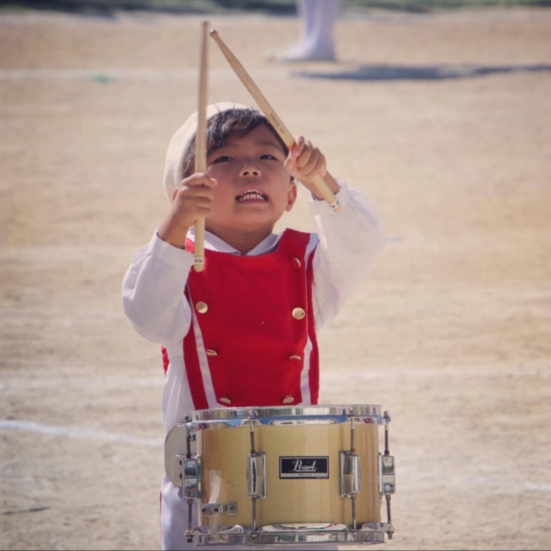 晴れた屋外のグラウンドで、赤と白のマーチングバンドの衣装を着た幼い男の子が、真剣な表情で小太鼓を演奏している。両手にスティックを持ち、口を少し開けて力強く太鼓を叩いている瞬間を捉えた写真。