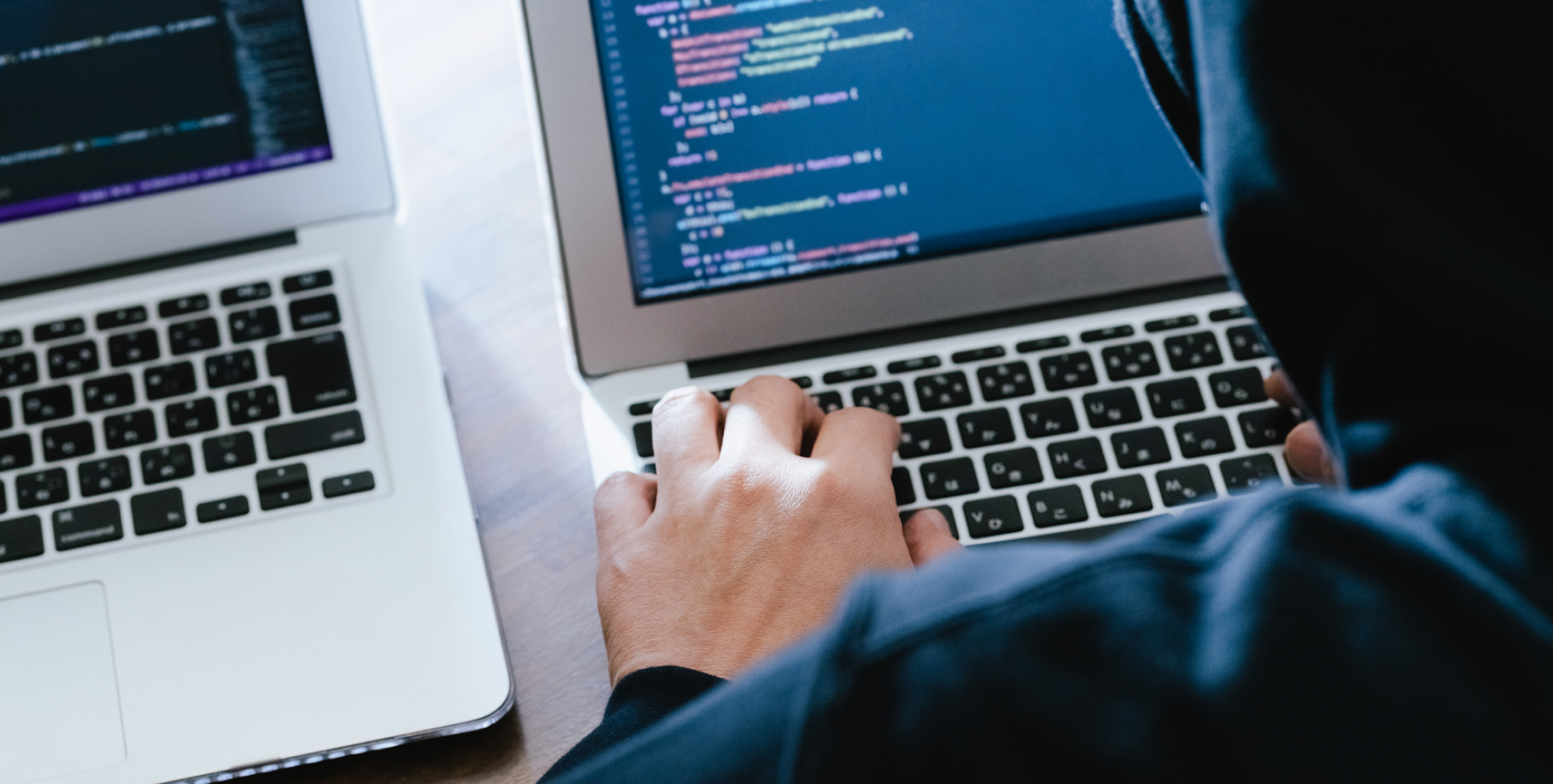 暗い環境で黒いパーカーを着た人物が、ノートパソコンに向かってプログラムコードを入力しているハッカーのイメージ