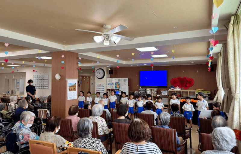 介護施設で大勢の利用者の方に見守られながら、年長さんが歌の発表をしている様子