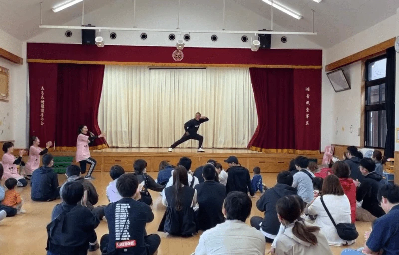 みもみ幼稚園のホールの壇上で男性の体操指導員がピースのポーズをしている様子