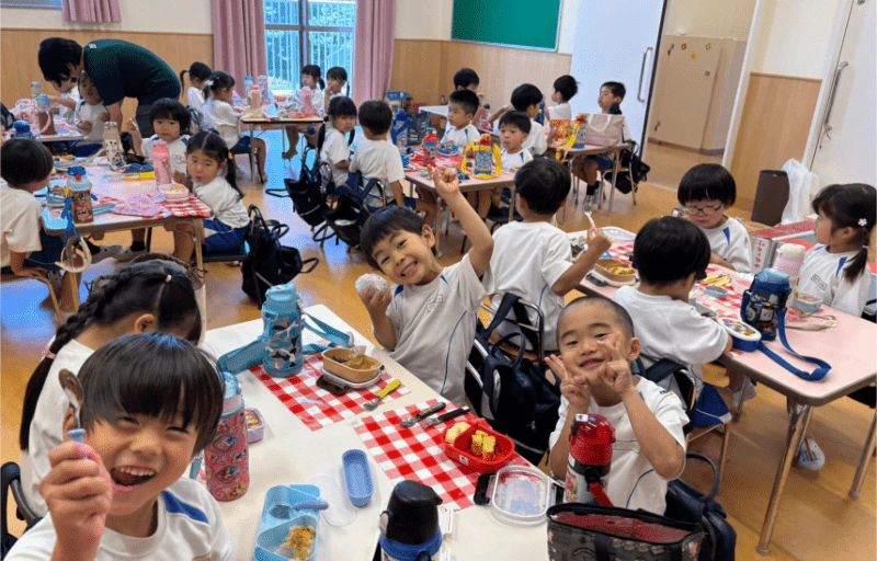 20名ほどの子ども達が机にお弁当を並べ笑顔で食べている様子