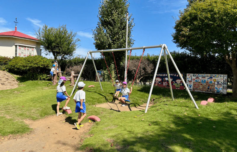複数の年長の女の子たちが青空の下楽しそうにブランコなどで遊んでいる様子