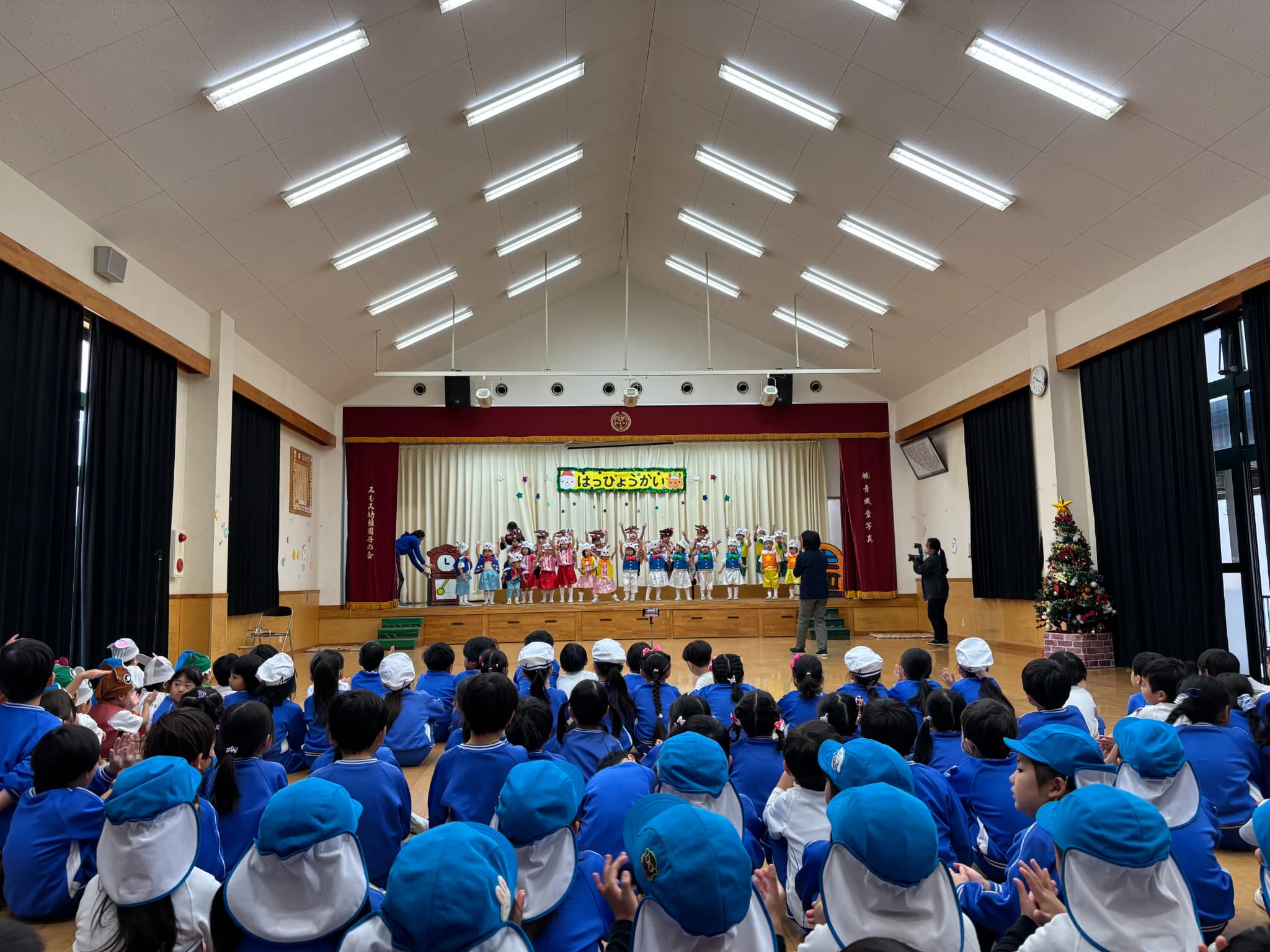 ホールで年少クラスが劇を発表しているところを他学年が見守っている様子